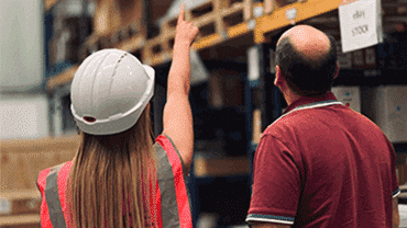 Woman in hard hat pointing to something in a warehouse, man standing next to woman looking at what she is pointing at